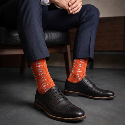 A person seated on a dark leather chair wearing bright orange crew socks with a repeating white origami pattern, navy dress pants, and black leather oxford shoes.