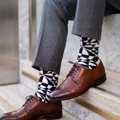 A close-up of a person's legs wearing brown leather oxford shoes, gray trousers, and black-and-white geometric patterned socks while sitting on stone steps.