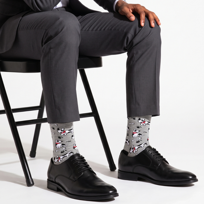 A man seated on a black chair wearing gray dress pants, black leather dress shoes, and light gray patterned crew socks with red and white illustrations.