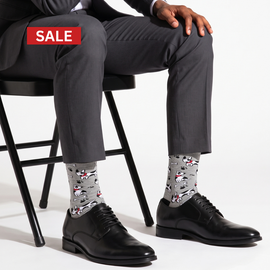 A man seated on a black chair wearing gray dress pants, black leather oxford shoes, and gray patterned socks with cartoon dog illustrations. A red 'SALE' banner appears in the top left on a white studio background.