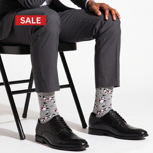 A man seated on a black chair wearing gray dress pants, black leather oxford shoes, and gray patterned socks with cartoon dog illustrations. A red 'SALE' banner appears in the top left on a white studio background.