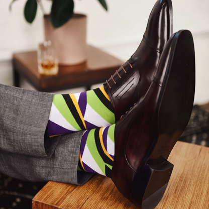 A person wearing gray trousers and dark brown oxford shoes with colorful striped socks, resting their legs on a wooden table in a styled indoor setting.