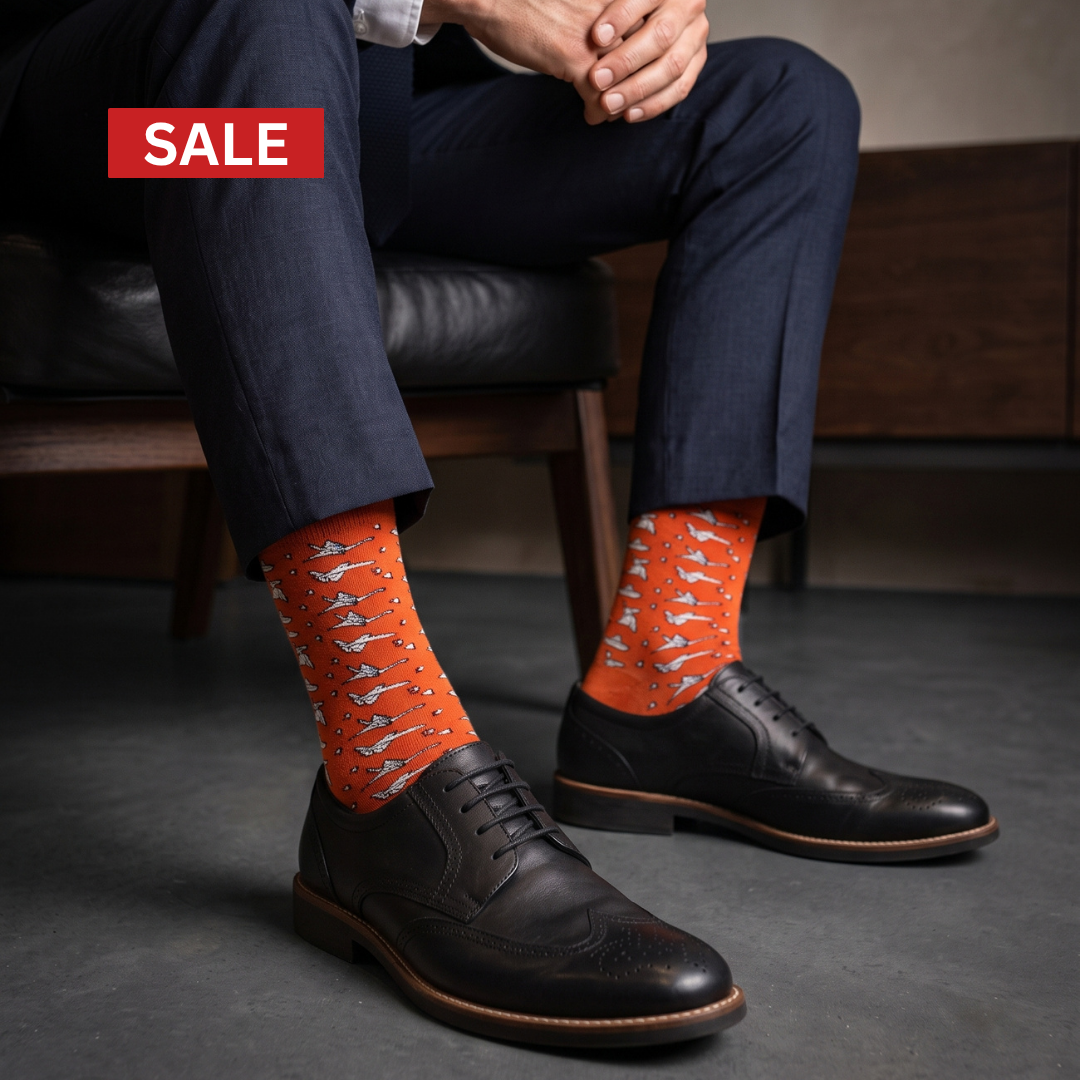 A person seated on a dark leather chair wearing bright orange crew socks with a white origami pattern, paired with dark navy trousers and black leather oxford shoes. A red 'SALE' banner appears in the top-left corner.