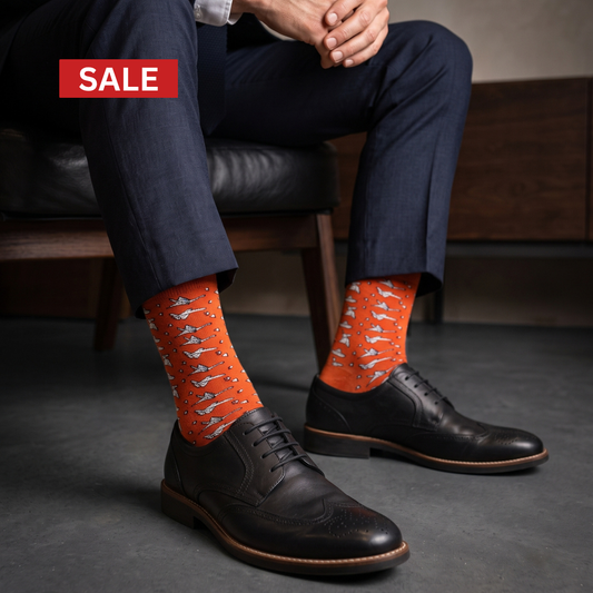 A person seated on a dark leather chair wearing bright orange crew socks with a white origami pattern, paired with dark navy trousers and black leather oxford shoes. A red 'SALE' banner appears in the top-left corner.